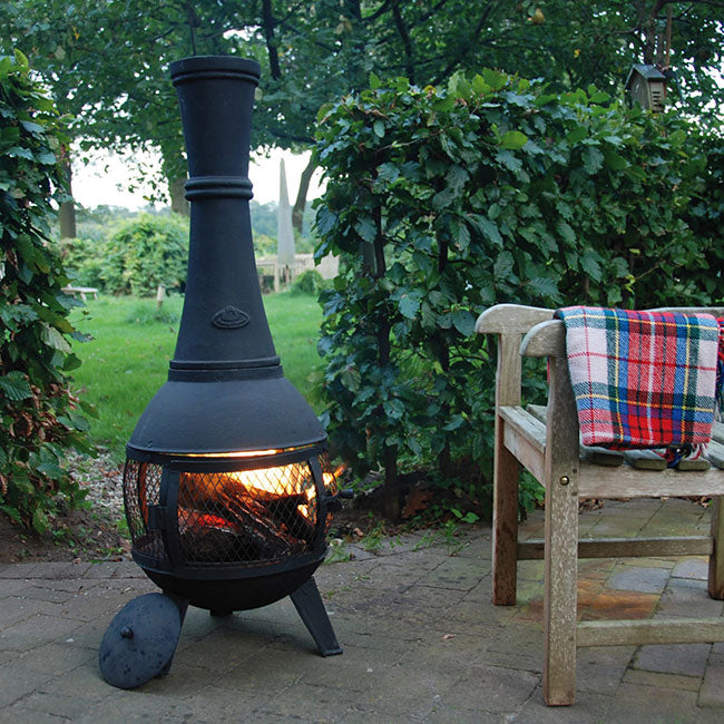 A cozy outdoor scene with a lit black chiminea on a patio. An old wooden bench with a colorful plaid blanket sits nearby, framed by lush green foliage.